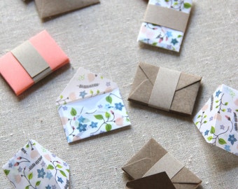 May include: A collection of eight small envelopes in various patterns and colors. The envelopes are made of paper and have a brown paper flap. Some of the envelopes have a floral pattern, while others are solid pink or brown.