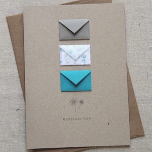 May include: A brown card with three envelopes on it. The top envelope is gray, the middle envelope is white with a blue floral pattern, and the bottom envelope is blue. The card says "Missing You" and has a small drawing of two dandelions.