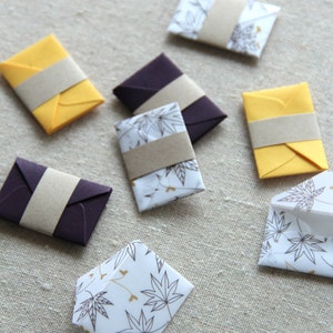 May include: A collection of small, colorful envelopes with different patterns and designs. Some envelopes are yellow, some are purple, and some are white with a leaf pattern. The envelopes are all closed and have a brown paper strip across the front.