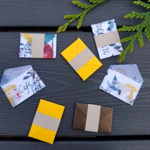 May include: Eight small envelopes with different designs and colors. Some envelopes have a brown paper flap, while others have a yellow paper flap. The envelopes are on a dark gray wooden surface.