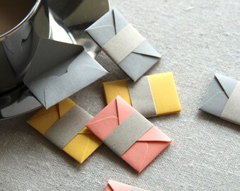 May include: A collection of small, folded envelopes in various colors, including gray, yellow, and pink. Each envelope has a gray paper strip across the front.