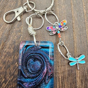 Peut inclure: Plusieurs porte-clés avec des attaches argentées. L'un d'eux présente un pendentif rectangulaire avec un motif tourbillonnant bleu, noir et violet. Deux breloques libellules, une avec des pierres précieuses colorées et l'autre turquoise, pendent des anneaux.