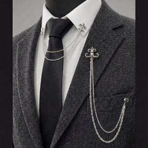 May include: A silver-toned metal collar pin featuring two fleur-de-lis emblems connected by double chains, paired with a matching lapel brooch that has a decorative crest and two long dangling chains attached to a small circular stud on the jacket.