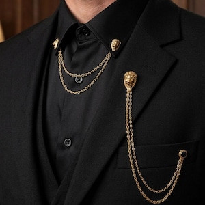 May include: A black suit with a black button-down shirt and a gold lion-head chain accessory. The chain drapes from the collar and lapel, adding a touch of elegance. The suit has a classic, tailored fit.