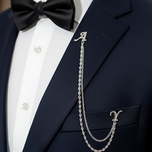 May include: A navy blue suit with a black bow tie and a silver chain brooch. The brooch features the letter "A" at the top and the letter "Y" at the bottom, connected by a delicate chain. The suit has a white dress shirt.