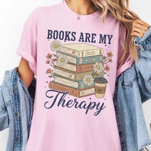May include: Pink t-shirt with the text "BOOKS ARE MY Therapy" and a graphic of stacked books, daisies, and a coffee cup. The shirt is worn with a denim jacket.