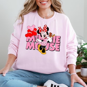 A woman wearing a pink sweatshirt with a Minnie Mouse graphic and the text "Minnie Mouse" is sitting in a room with plants and a glass globe.