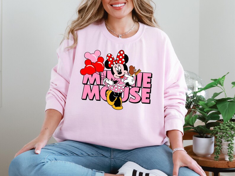 A woman wearing a pink sweatshirt with a Minnie Mouse graphic and the text "Minnie Mouse" is sitting in a room with plants and a glass globe.