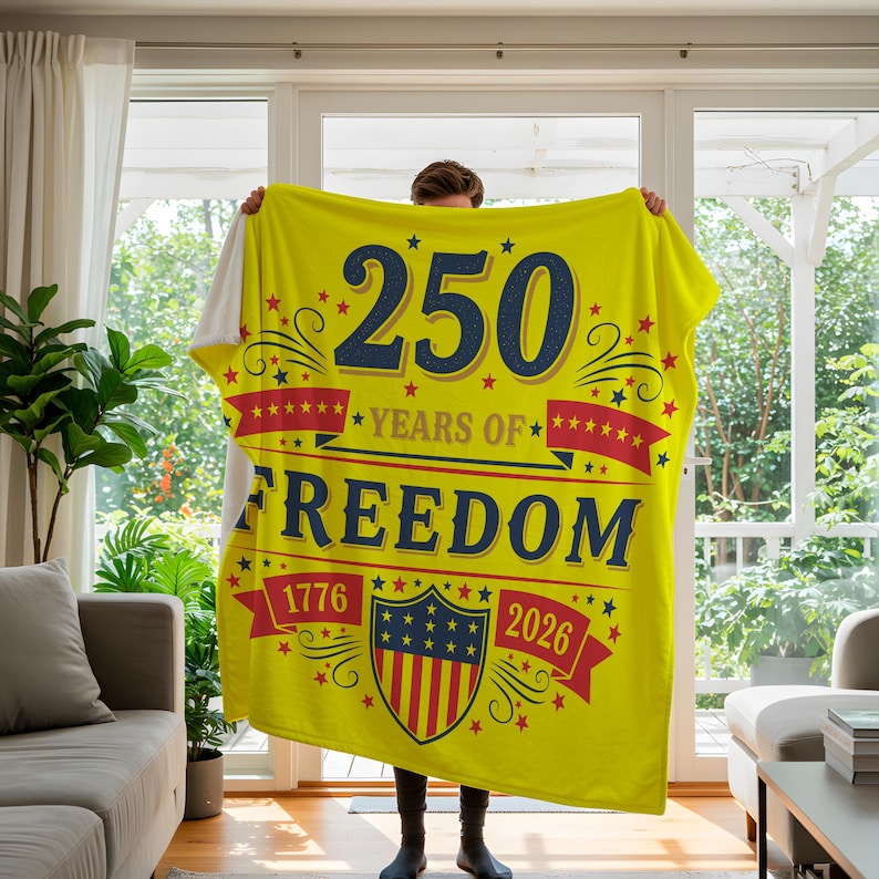 A person is holding up a large yellow blanket with the text "250 YEARS OF FREEDOM" and "1776 2026" printed on it.