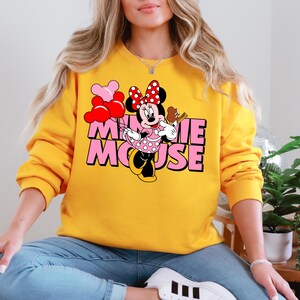 A woman wearing a yellow sweatshirt with a Minnie Mouse graphic on it, sitting in a relaxed position.