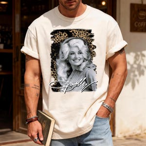 May include: A cream-coloured short-sleeve t-shirt featuring a black and white portrait of Dolly Parton centred within a leopard print graphic background. The name Dolly is written in a white cursive script font across the bottom of the graphic.