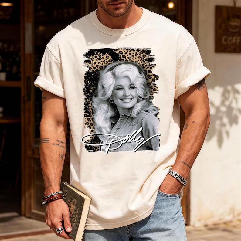 May include: A cream-coloured short-sleeve t-shirt featuring a black and white portrait of Dolly Parton centred within a leopard print graphic background. The name Dolly is written in a white cursive script font across the bottom of the graphic.