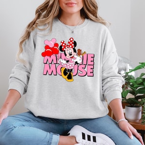 A woman wearing a gray sweatshirt with a Minnie Mouse graphic and the text "Minnie Mouse" is sitting on the floor in a room with potted plants.