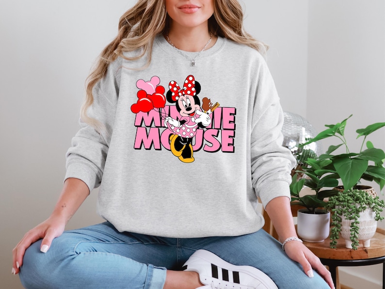 A woman wearing a gray sweatshirt with a Minnie Mouse graphic and the text "Minnie Mouse" is sitting on the floor in a room with potted plants.