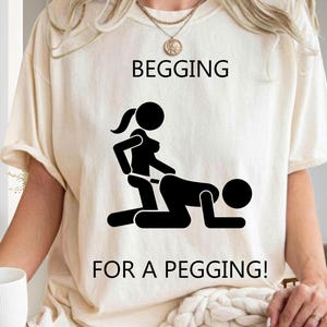 May include: A cream-colored t-shirt featuring a black graphic of two stick figures in a sexual position. The text "BEGGING" is printed above the graphic, and the text "FOR A PEGGING!" is printed below the graphic in a simple black font.