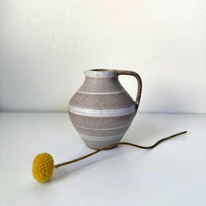 May include: A small, gray ceramic pitcher with horizontal white stripes and a brown handle. A yellow, spherical flower head on a thin stem rests in front of the pitcher. The pitcher is on a white surface.