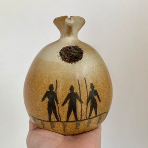 May include: A ceramic pitcher with a brown glaze and a design of three figures in black silhouette. The figures are holding staffs and are standing on a line.