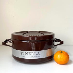 May include: A brown Finella Wartsila pot with a lid and handles. The pot has a silver band with the Finella logo and "Made in Finland" text. A small orange tangerine sits next to the pot.