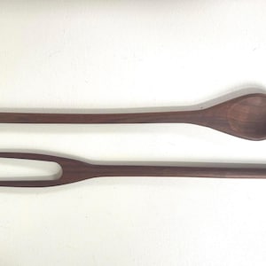 May include: A wooden salad spoon and fork set. The spoon has a rounded bowl and long handle. The fork has two tines and a long handle. Both are made of dark brown wood.