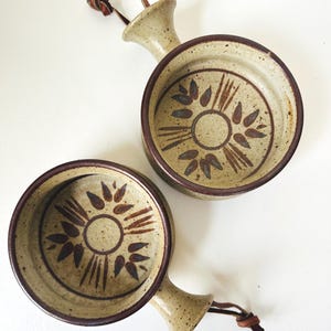 May include: Two round ceramic bowls with handles, featuring a speckled beige finish and a dark brown rim. Each bowl displays a central circular motif surrounded by dark brown leaf-like patterns. A brown leather cord is tied to each handle.