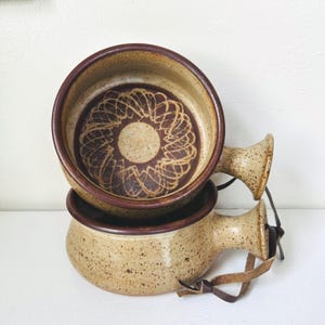 May include: Two stacked ceramic bowls with handles, featuring a speckled beige finish and a dark brown rim. The interior of the top bowl displays a circular, intricate brown line pattern. A brown leather cord is attached to the handle of the bottom bowl.