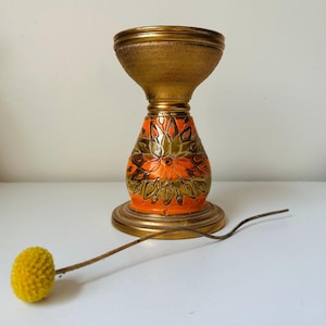 May include: A vintage candle holder with a gold-colored top and base, and a patterned orange and green center. A yellow flower with a long stem rests nearby. The candle holder is likely made of ceramic or metal.