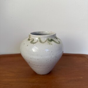 May include: A small, white ceramic vase with a green glaze design around the rim. The vase has a rounded body and a slightly flared opening.