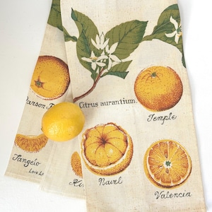May include: A folded beige kitchen towel with illustrations of oranges and citrus blossoms. The towel features the names of different orange varieties, including "Temple" and "Valencia". A yellow lemon rests on the towel.