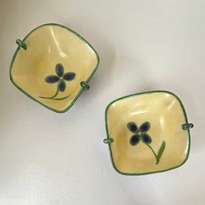 May include: Two square, yellow ceramic bowls with green rims and handles. Each bowl features a blue flower design with green stems. The bowls are set against a white background.