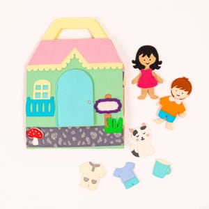 Op de afbeelding: Een vilt speelset met een huis, twee figuren, een kat en accessoires. Het huis is groen met een blauwe deur en een roze dak. De figuren zijn een meisje met bruin haar dat een roze jurk draagt en een jongen met bruin haar dat een oranje shirt en een blauwe broek draagt. De kat is wit met zwarte vlekken. De accessoires omvatten een paddenstoel, een bord, een kopje en twee kledingstukken.