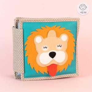 Könnte beinhalten: Ein weiches Stoffbuch für Babys und Kleinkinder mit einem Löwen mit einer leuchtend orangefarbenen Mähne, einem weißen Gesicht und einer roten Zunge. Das Buch hat einen grauen und weißen Polka-Dot-Umschlag und den Text '+12 M' auf der Vorderseite.