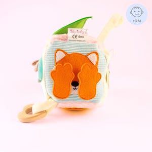 Puede incluir: Cubo de actividades para bebés con cara de zorro y patas naranjas que cubren los ojos. El cubo tiene un anillo de madera, una hoja verde y una etiqueta que dice "Jolly Designs CE 6m+". Juguete para bebés de 6 meses en adelante, con diferentes texturas.