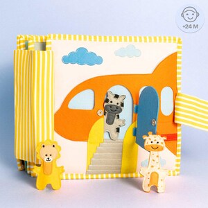 Könnte beinhalten: Ein gelbes und oranges Stoffbuch mit einem Flugzeug, einem Zebra, einer Giraffe und einem Löwen. Das Buch hat eine Klappe, die sich öffnet, um die Figuren zu enthüllen. Das Buch ist für Kinder ab 24 Monaten konzipiert.