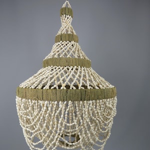 Può includere: Una lampada a sospensione con una struttura a livelli avvolta in spago verde oliva, decorata con una rete complessa e fili a cascata di piccole conchiglie bianche. Il design include un gancio metallico sulla parte superiore.
