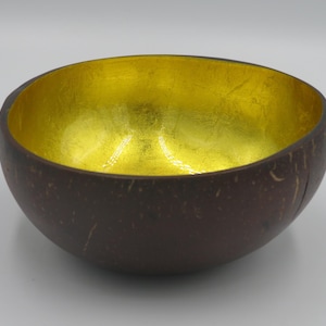 Op de afbeelding: Een ronde kom gemaakt van een donkerbruine kokosnootschaal met een getextureerde, natuurlijke buitenkant. De binnenkant van de kom heeft een gladde, reflecterende metallic bladgouden afwerking die contrasteert met het organische, donkerbruine oppervlak.