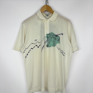 May include: Cream-colored polo shirt with a three-button placket and short sleeves. The shirt features a graphic design with a mountain-like pattern in black and teal. The Adidas logo is visible on the left chest.