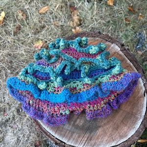 May include: A colorful, handmade crochet hat with a ruffled brim. The hat features a mix of blue, purple, green, and pink yarn. The hat is sitting on a wooden surface.
