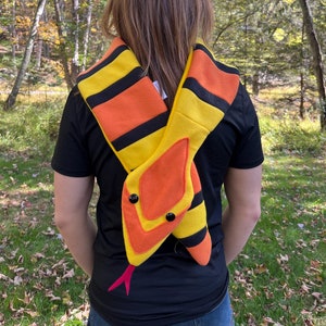 Könnte beinhalten: Ein gelb-orange-schwarzer Schal in Schlangenform mit schwarzen Knopfaugen und einer rosa Zunge. Der Schal wird um Hals und Schultern getragen. Der Hintergrund ist eine natürliche Umgebung im Freien mit Bäumen.