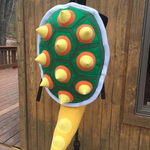 Puede incluir: Un disfraz de caparazón de tortuga verde, amarillo y naranja con púas. El caparazón está unido a una cola amarilla.