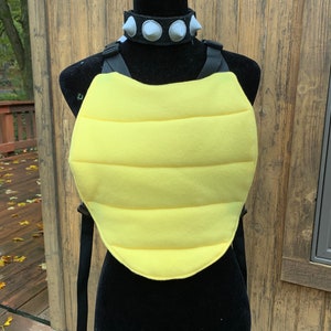 May include: A yellow, padded costume piece with black straps and a black spiked collar. The costume piece is designed to resemble a turtle shell.