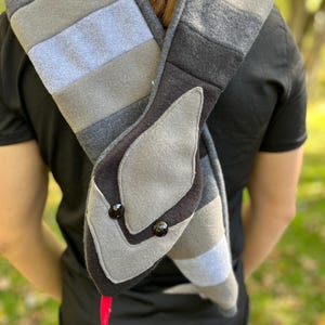 May include: A gray and black snake-shaped scarf with a red tongue and black button eyes. The scarf is made of various shades of gray fabric and is draped over a person's shoulders.