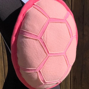 May include: Pink plush turtle shell costume accessory with a white strap. The shell has a textured design with raised lines.