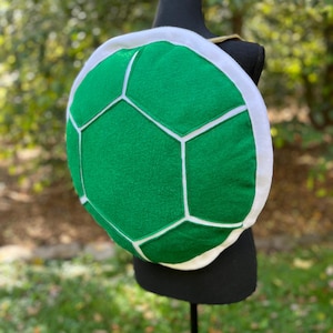 May include: A green turtle shell costume with white trim and white segmented lines. The shell is displayed on a black mannequin. The costume is made of soft material and is designed to be worn on the back.