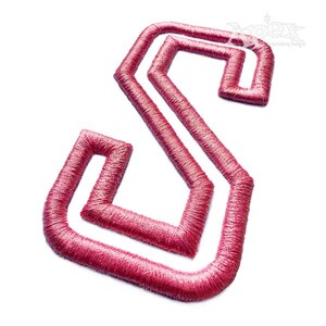 May include: A pink embroidered letter S with a bold, geometric design. The letter is outlined in white and has a double-stitched effect.