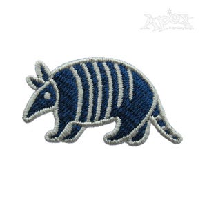 Puede incluir: Parche bordado de un armadillo azul con rayas blancas. El armadillo está mirando hacia la izquierda y tiene un ojo blanco.