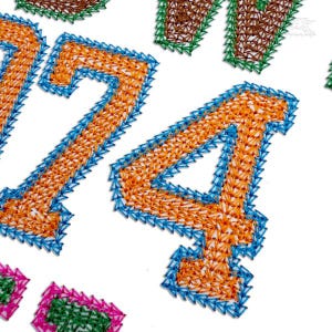 May include: Close-up of embroidered numbers '24' in orange with blue trim, and other numbers in green and brown. The numbers are stitched on a white background. The style is reminiscent of a sports jersey.