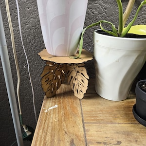 Könnte beinhalten: Ein hölzerner Pflanzenständer mit Blättermuster. Der Ständer hat eine runde Oberseite und drei blattförmige Beine. Ein rosa-weiß gemusterter Blumentopf steht auf dem Ständer, daneben eine grüne Pflanze. Der Ständer steht auf einer Holzoberfläche.