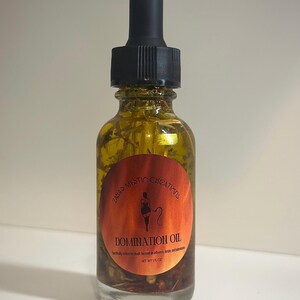 May include: A clear glass bottle of Domination Oil with a black dropper. The bottle has a round, orange label with the text "Zaba's Mystic Creations" and "DOMINATION OIL". The oil contains herbs and is 1 fl oz.