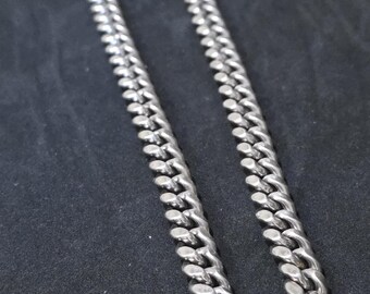 May include: A silver-colored curb chain necklace. The chain is made of interlocking oval links, creating a bold and classic design. The necklace is displayed against a dark background, highlighting its metallic sheen. A simple, yet stylish piece of jewelry.