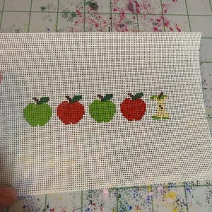 May include: A cross-stitch canvas featuring five apples. Two green and two red apples are whole, while the last is a core. The apples have green leaves and are stitched on a white mesh background. The canvas is rectangular.
